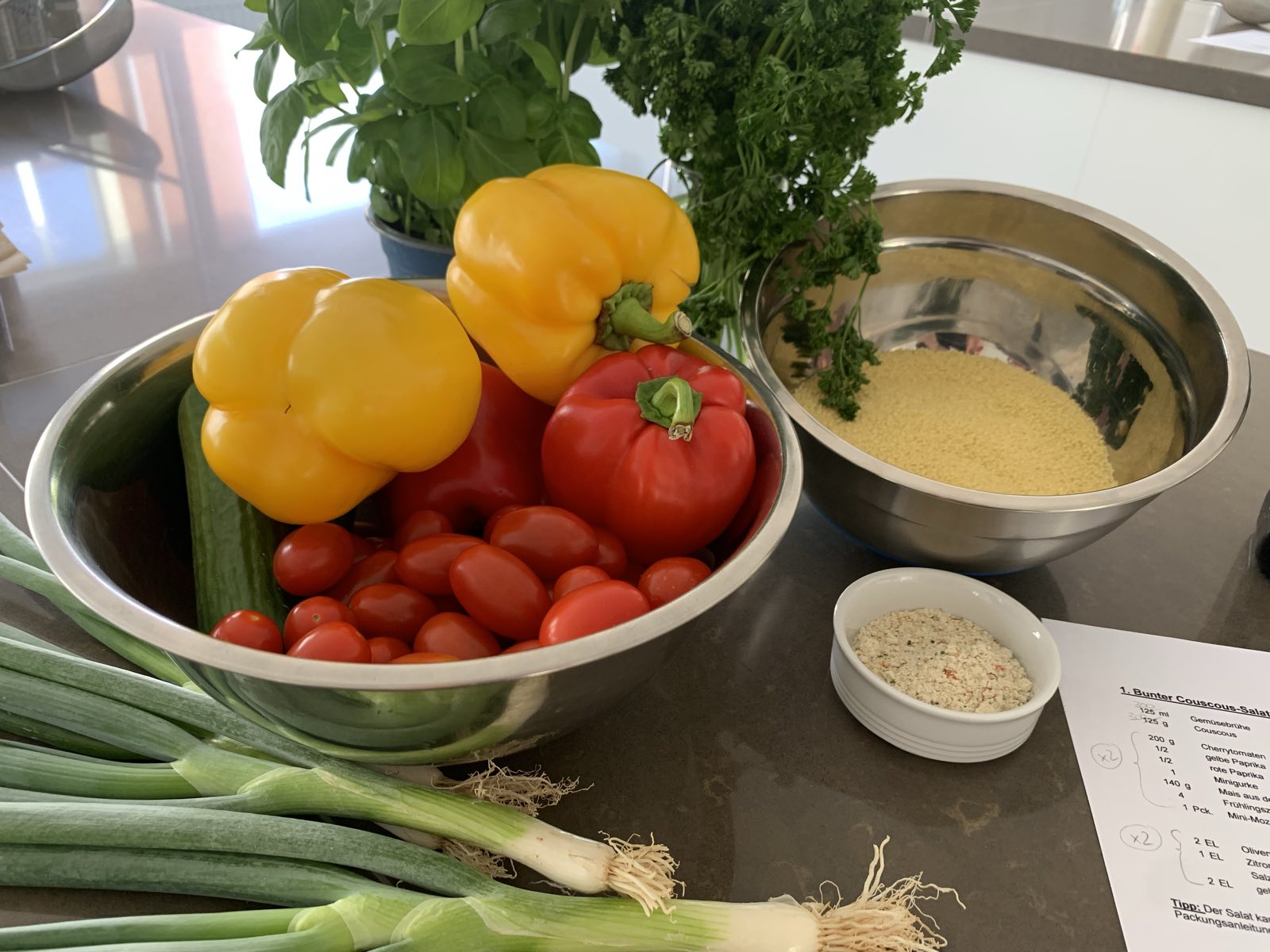 Vorbereiteter Arbeitsplatz für einen Kochworkshop: eine Schüssel mit gelben und roten Paprika, Tomaten und Gurke, daneben Couscous, Frühlingszwiebeln, frischer Basilikum und Petersilie sowie ein ausgedrucktes Rezept