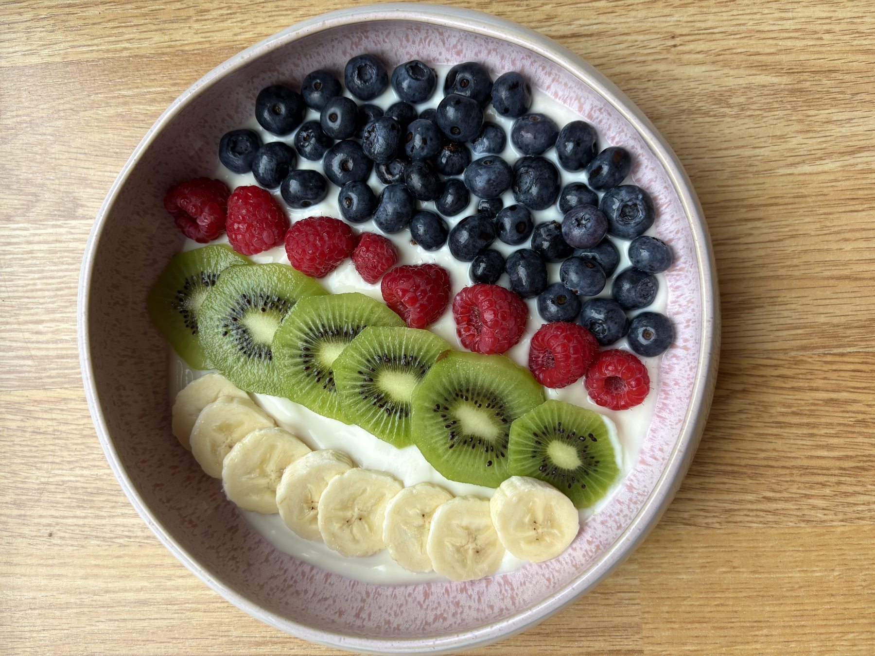 Schale mit Joghurt, Banane, Kiwi, Himbeeren und Heidelbeeren auf einem hellen Holztisch